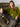 A woman with long brown hair sits confidently on a green velvet armchair, wearing the Sanctuary Clothing must have cardi martini olive, paired with dark pants and brown boots, her leg casually draped over the armrest.