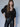 A woman with long brown hair and bangs models the Sanctuary Clothing meow cardi kitty camo leopard print cardigan with black faux leather pants and a black belt against a plain light gray background.