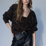 A woman with long brown hair and bangs models the Sanctuary Clothing meow cardi kitty camo leopard print cardigan with black faux leather pants and a black belt against a plain light gray background.