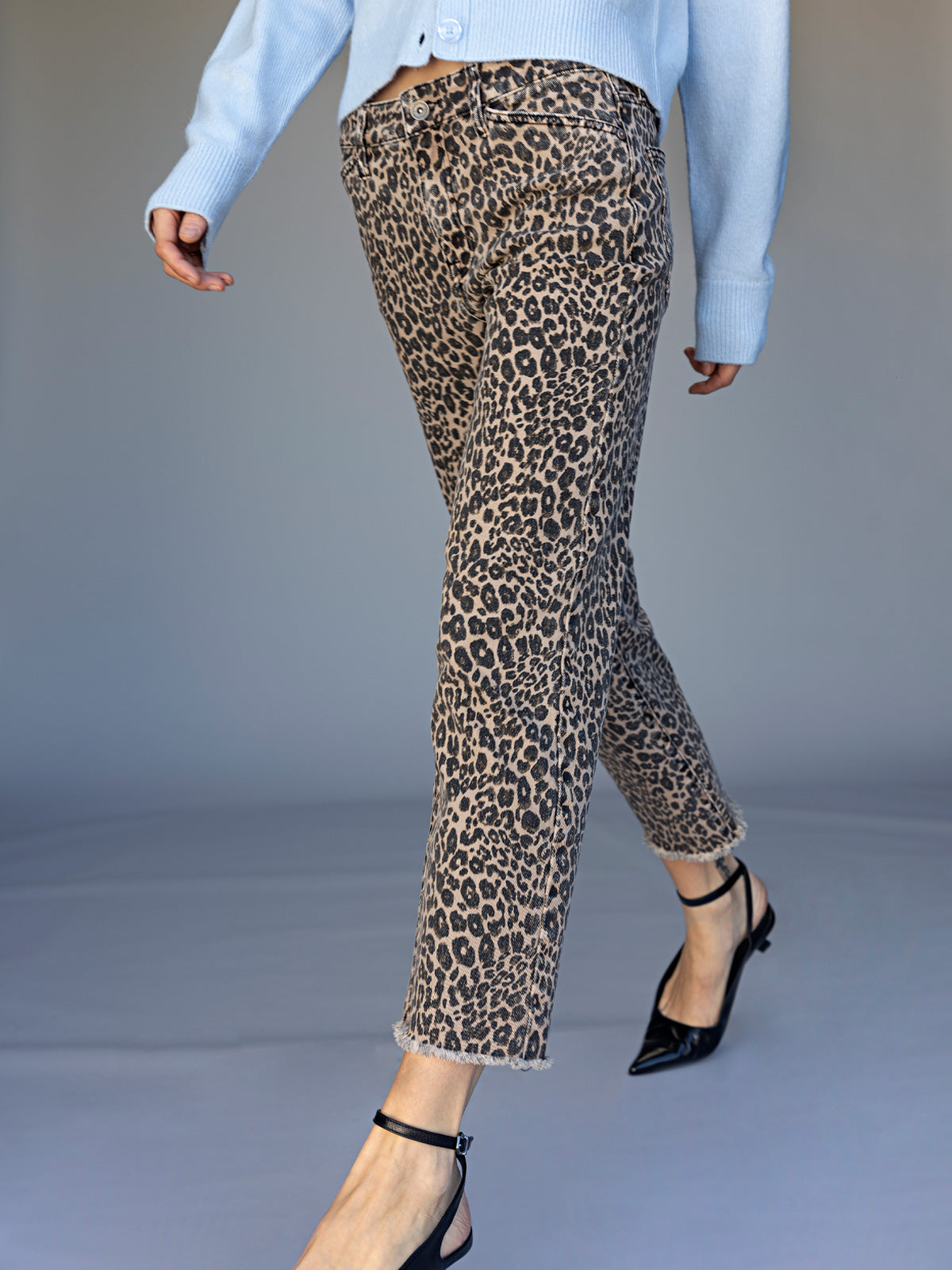 A person wearing Sanctuary Clothing cadence 90 straight crop jeans in french kitty leopard print, paired with a light blue sweater and black pointed-toe heels, walks against a plain background. Only the lower half is visible.