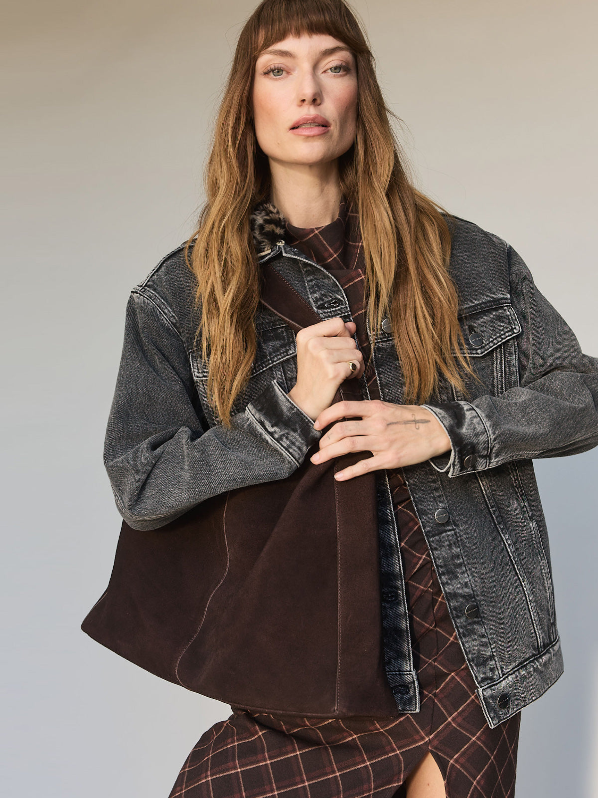 A woman with long brown hair wears a faded denim jacket over a patterned dress, holding the Sanctuary Clothing endless large hobo bag in chicory. She stands against a neutral background, looking confidently at the camera.