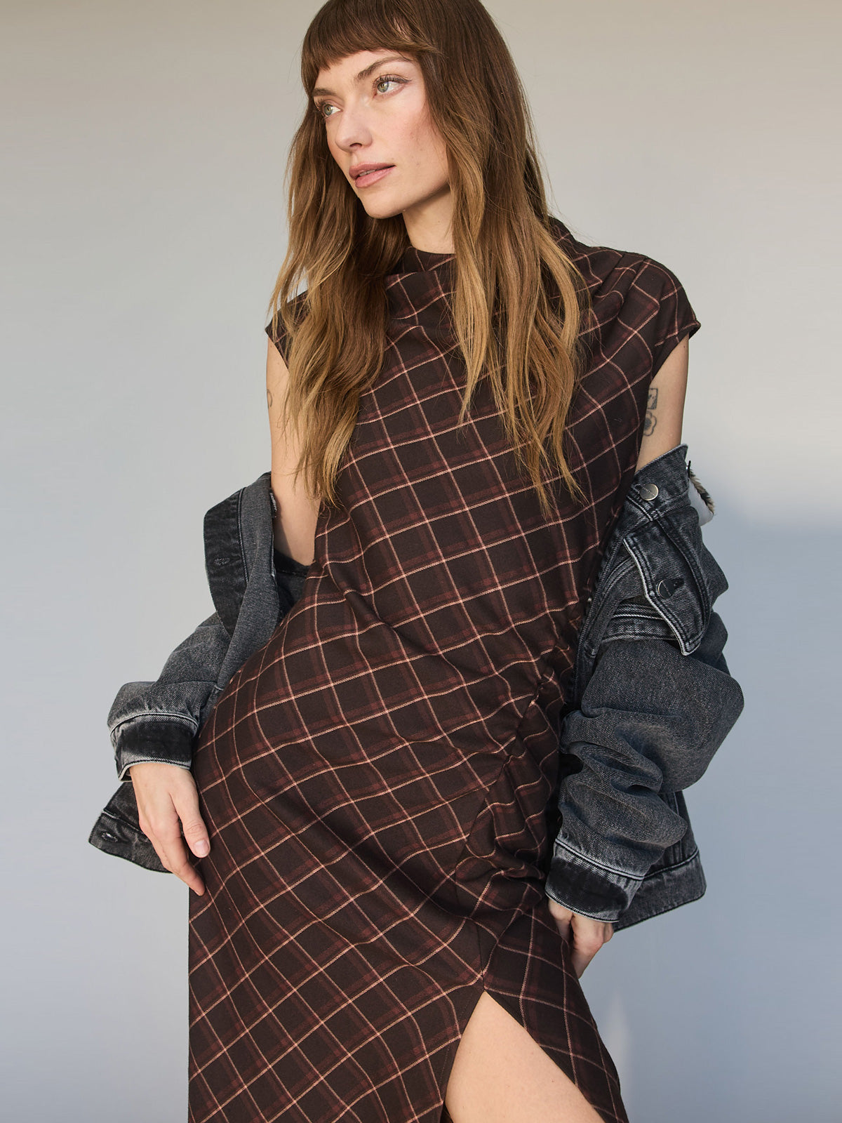A woman with long brown hair wears the Sanctuary Clothing autumns best sweater dress in cocoa plaid, featuring a thigh-high slit, styled with a black denim jacket over her shoulders, posing against a neutral background.