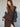 A woman with long brown hair wears the Sanctuary Clothing autumns best sweater dress in cocoa plaid, featuring a thigh-high slit, styled with a black denim jacket over her shoulders, posing against a neutral background.