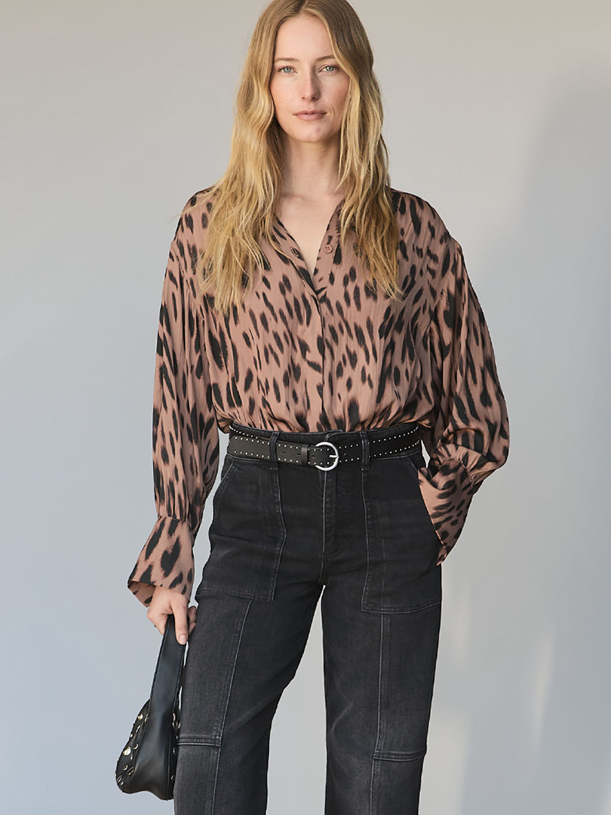 A woman with long blonde hair wears the Sanctuary Clothing "colette shirt mocha leo," a loose animal-print blouse, tucked into high-waisted dark trousers with white stitching, holding a black clutch against a light background.