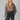 A woman with long blonde hair wears the Sanctuary Clothing "colette shirt mocha leo," a loose animal-print blouse, tucked into high-waisted dark trousers with white stitching, holding a black clutch against a light background.
