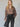 A woman with long blonde hair wears the Sanctuary Clothing "colette shirt mocha leo," a loose animal-print blouse, tucked into high-waisted dark trousers with white stitching, holding a black clutch against a light background.