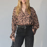 A woman with long blonde hair wears the Sanctuary Clothing "colette shirt mocha leo," a loose animal-print blouse, tucked into high-waisted dark trousers with white stitching, holding a black clutch against a light background.