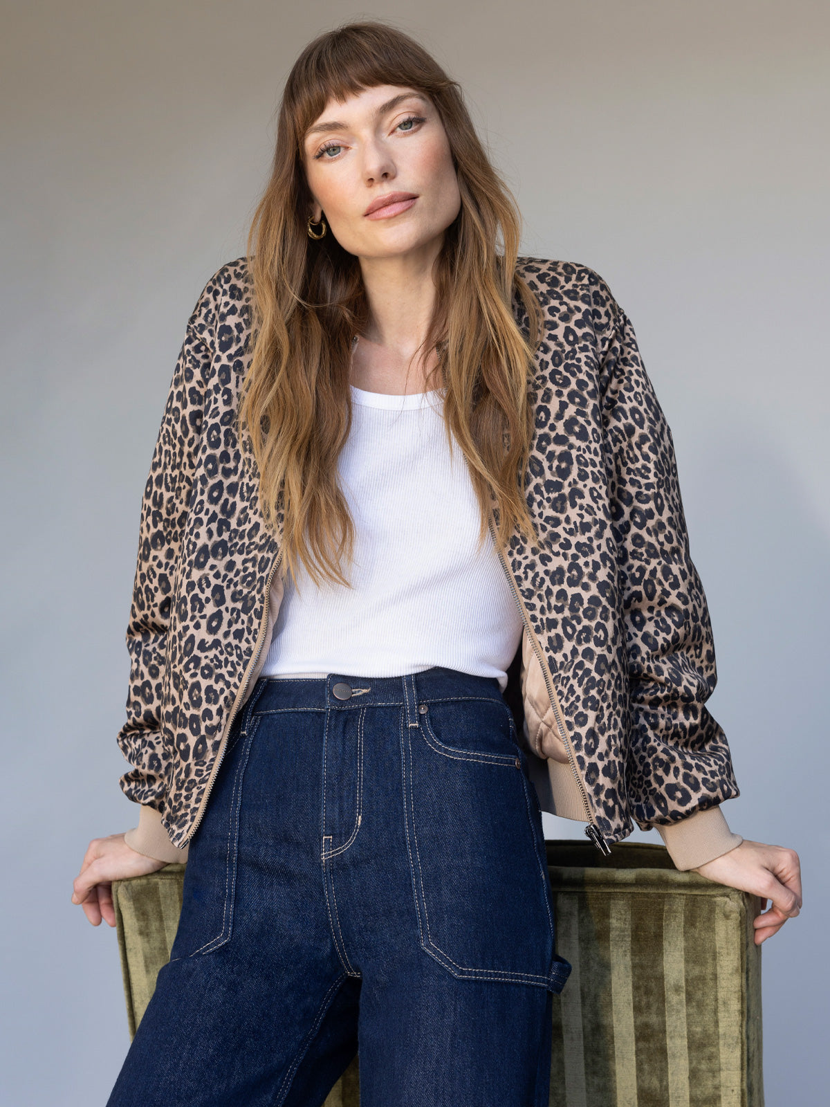 A woman with long brown hair and bangs wears the Monroe Sateen Bomber Jacket French Kitty by Sanctuary Clothing over a white top and high-waisted blue jeans, leaning on a green chair against a neutral background.