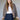 A woman with long brown hair and bangs wears the Monroe Sateen Bomber Jacket French Kitty by Sanctuary Clothing over a white top and high-waisted blue jeans, leaning on a green chair against a neutral background.