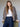 A woman with long brown hair and bangs wears the Monroe Sateen Bomber Jacket French Kitty by Sanctuary Clothing over a white top and high-waisted blue jeans, leaning on a green chair against a neutral background.