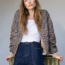 A woman with long brown hair and bangs wears the Monroe Sateen Bomber Jacket French Kitty by Sanctuary Clothing over a white top and high-waisted blue jeans, leaning on a green chair against a neutral background.