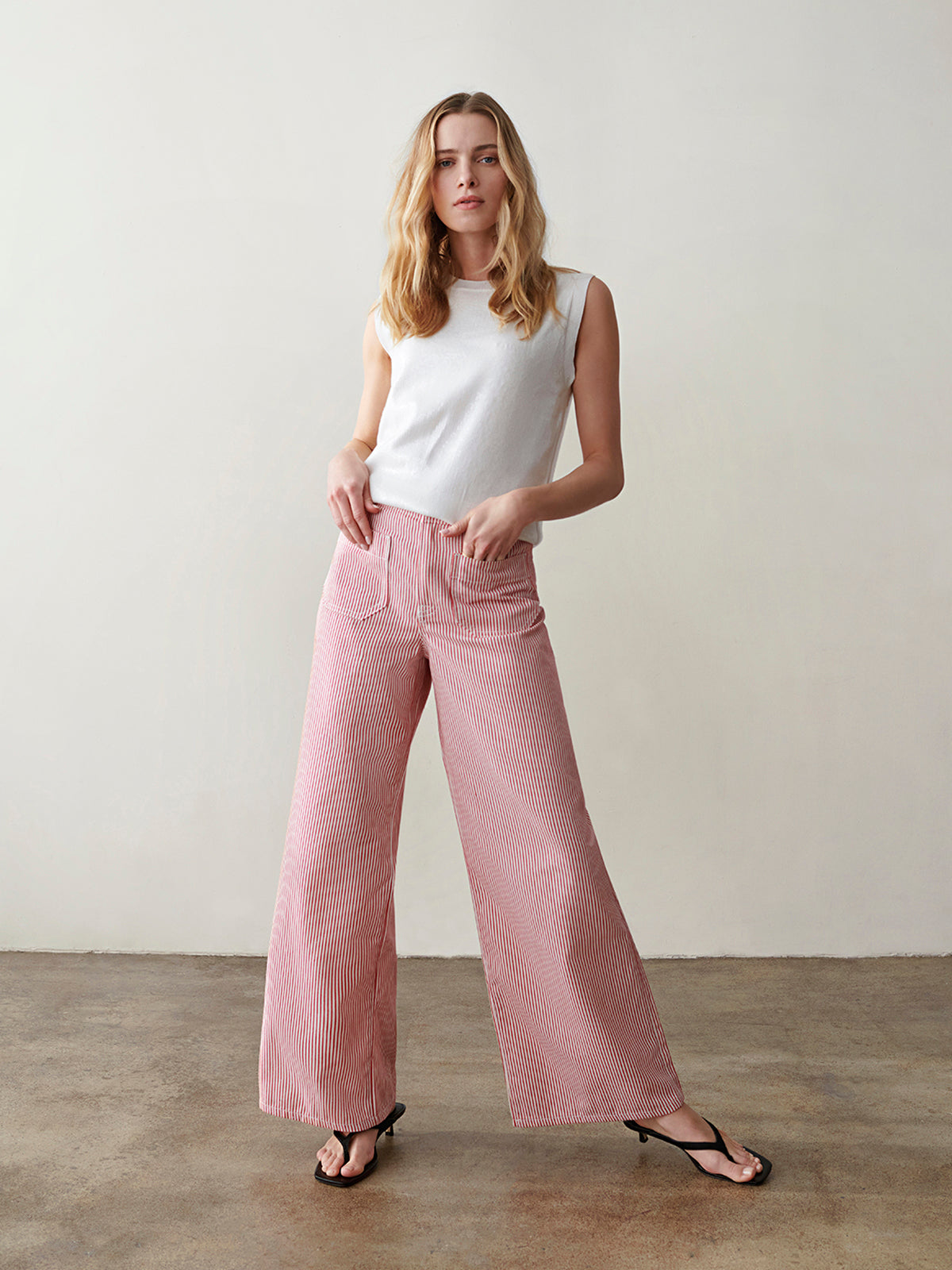 A woman with long blonde hair wears the Sanctuary Clothing marine full length semi high rise pant in galley stripe and a sleeveless white top, standing on a polished concrete floor against a plain light-colored wall.