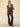 A woman stands against a beige background, wearing a brown blazer, white shirt, dark blue jeans, black ankle boots, and carrying Sanctuary Clothing’s perfect bucket bag leopard over her shoulder as she looks confidently at the camera.
