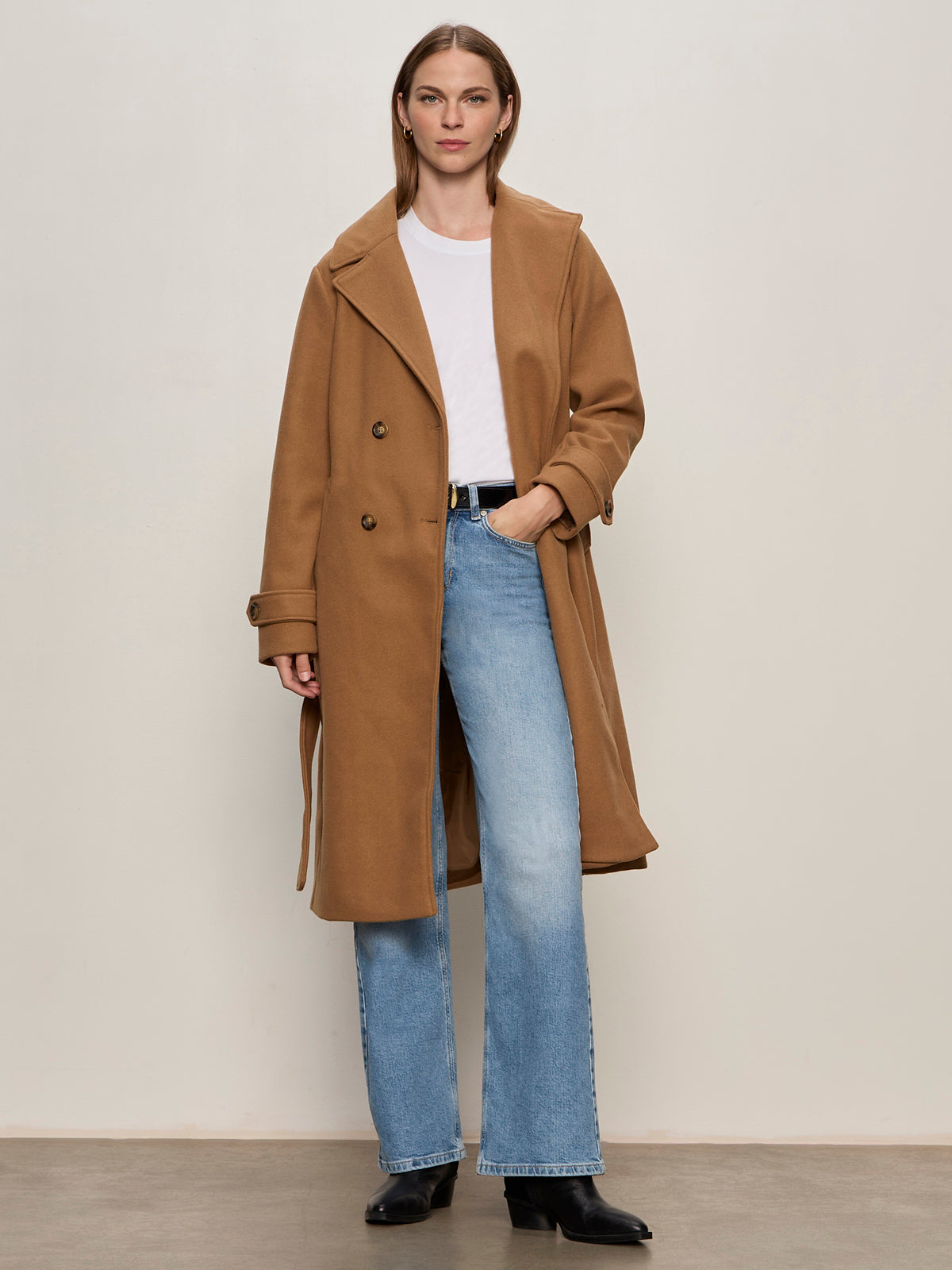 A woman with straight, shoulder-length hair stands against a plain background wearing the Sanctuary Clothing Aria Wrap Coat in camel over a white tee, light blue jeans, and black boots, with her left hand in her pocket.
