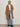 A woman with straight, shoulder-length hair stands against a plain background wearing the Sanctuary Clothing Aria Wrap Coat in camel over a white tee, light blue jeans, and black boots, with her left hand in her pocket.