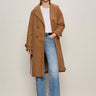 A woman with straight, shoulder-length hair stands against a plain background wearing the Sanctuary Clothing Aria Wrap Coat in camel over a white tee, light blue jeans, and black boots, with her left hand in her pocket.