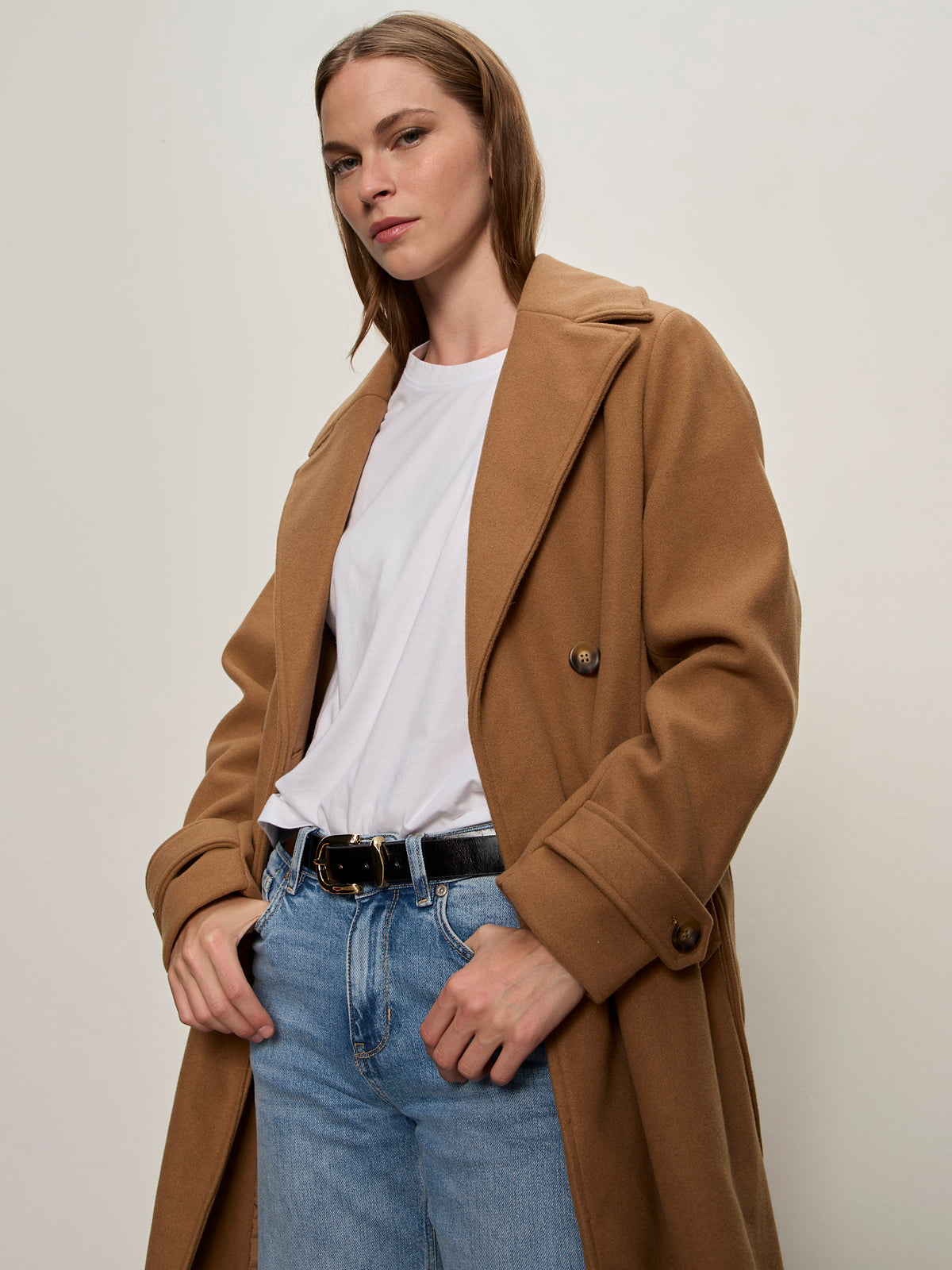 A woman stands with her hands in her pockets against a plain light background, wearing the Aria Wrap Coat Camel by Sanctuary Clothing over a white t-shirt and light blue jeans.