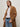 A woman stands with her hands in her pockets against a plain light background, wearing the Aria Wrap Coat Camel by Sanctuary Clothing over a white t-shirt and light blue jeans.