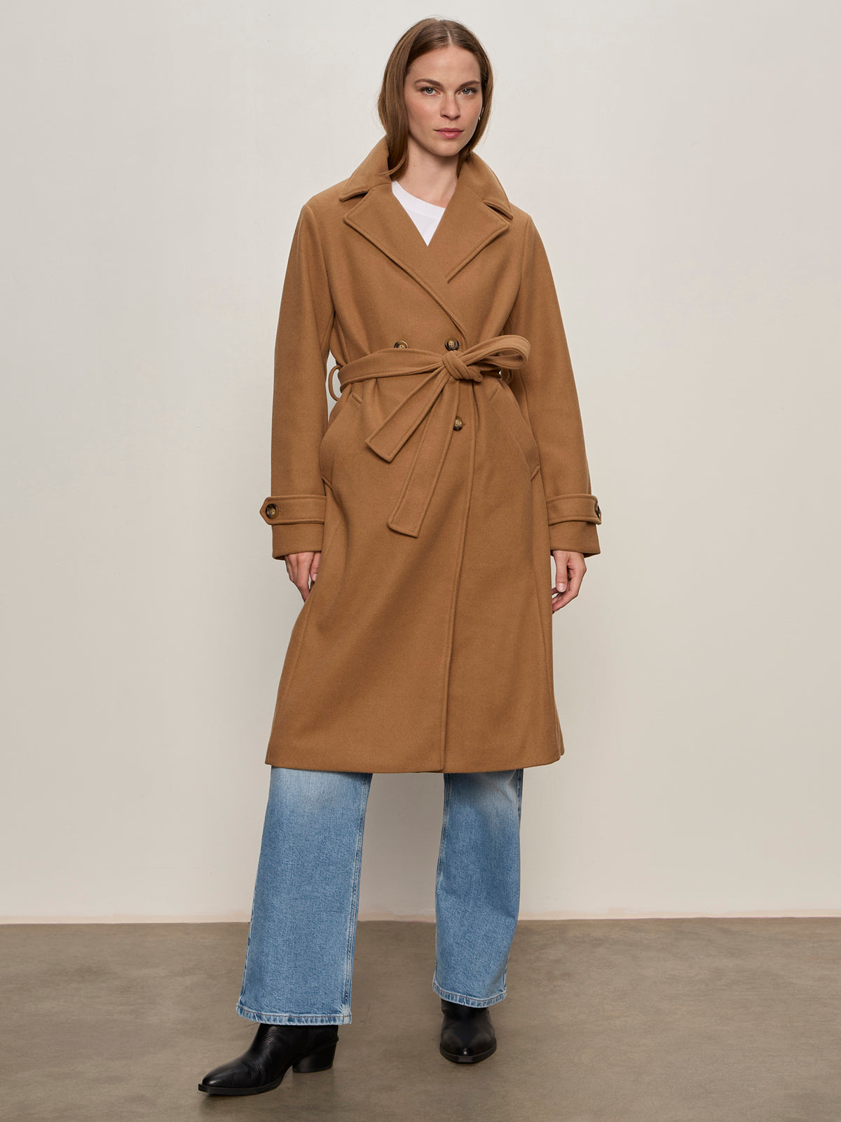 A woman with straight, shoulder-length hair and a neutral expression stands against a plain background wearing the Sanctuary Clothing Aria Wrap Coat in camel over light blue jeans and black ankle boots.