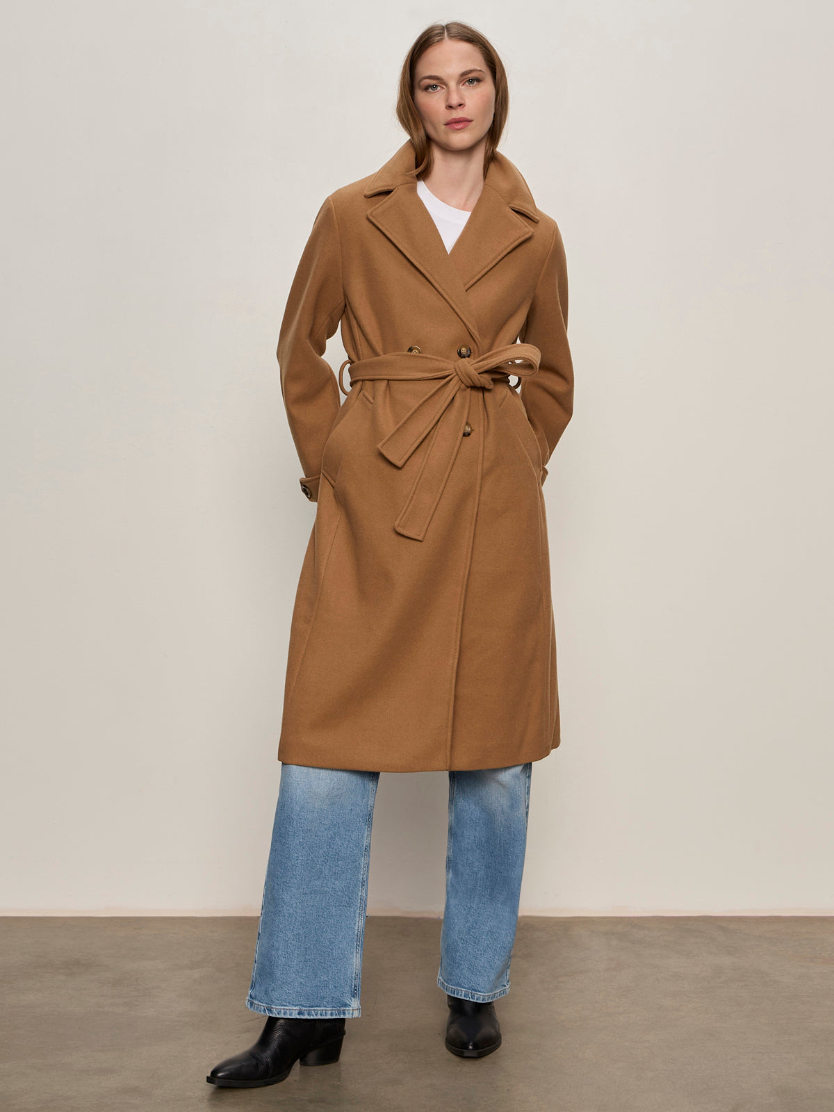 A woman stands against a plain background, wearing the Aria Wrap Coat Camel by Sanctuary Clothing over a white top, blue jeans, and black ankle boots, with her hands in her pockets.