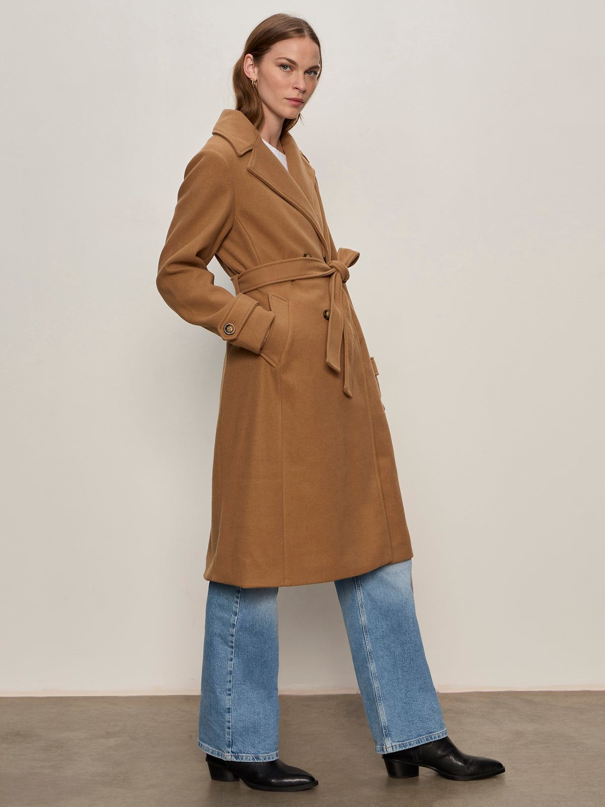 A woman with straight, shoulder-length hair and a neutral expression stands against a light wall, wearing the Sanctuary Clothing Aria Wrap Coat in camel over light blue jeans and black boots.