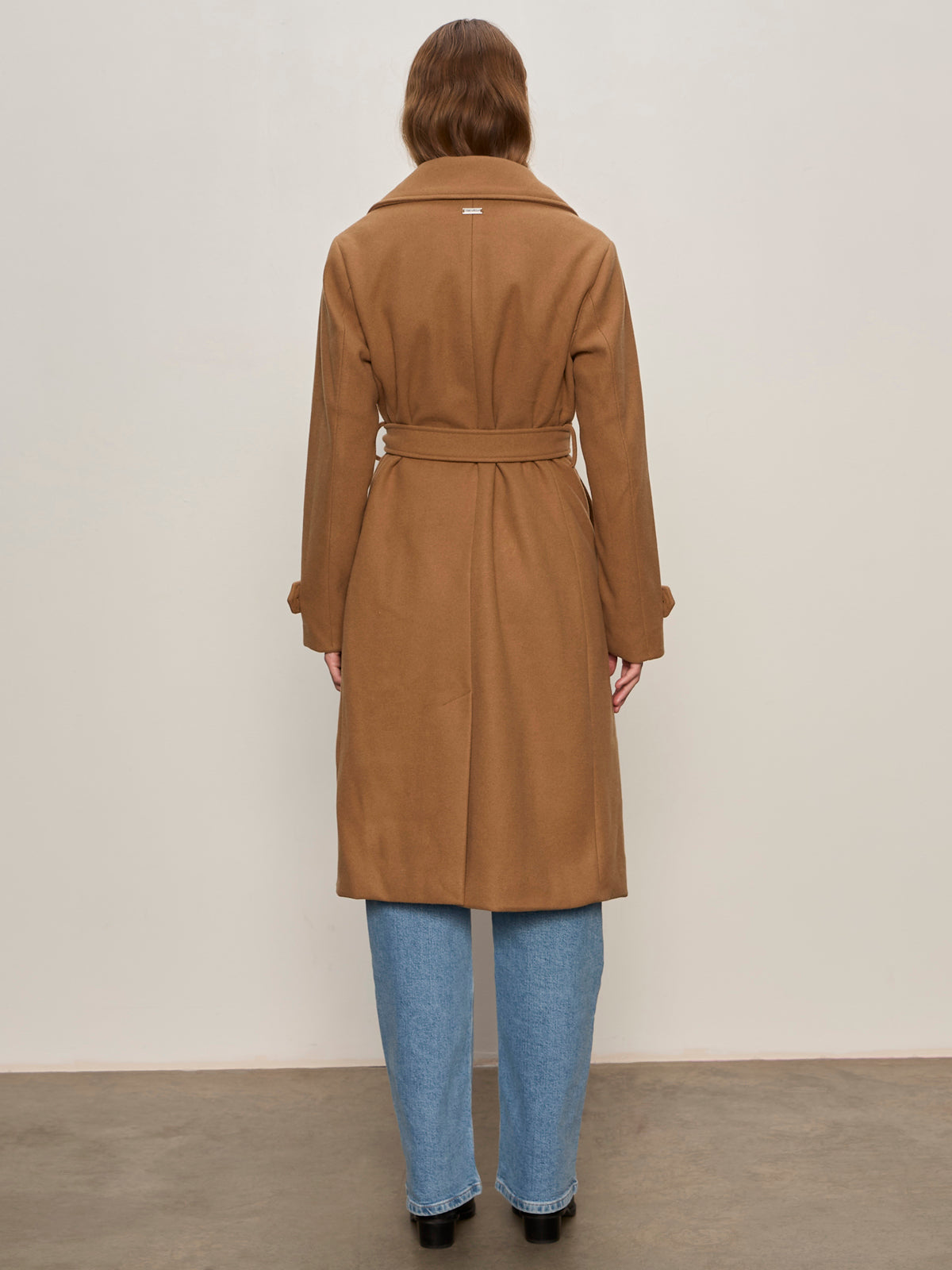A person with long hair stands facing away against a beige background, wearing the Sanctuary Clothing Aria Wrap Coat in camel over light blue jeans and black shoes.