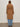 A person with long hair stands facing away against a beige background, wearing the Sanctuary Clothing Aria Wrap Coat in camel over light blue jeans and black shoes.