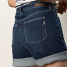 A person in Sanctuary Clothing's cuffed denim short dark with a leather patch and visible back pockets, paired with a striped top, stands in profile to showcase the side and back of their outfit.