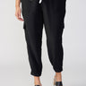 A person is wearing The Harmony Black cargo pants with elasticated cuffs from Sanctuary Clothing, standing with one hand in their pocket. They are also wearing black slide sandals featuring block heels and a white top tied at the waist. The background is plain and neutral.